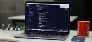 MacBook Pro displaying source code in VS Code with a red Nescafé mug, smartphone, and LEGO figures on a white desk in the background.