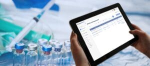 Hands holding a tablet with a pharmaceutical waste management dashboard, with medical vials, syringe, and lab equipment in background.